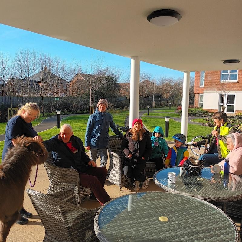 a group of people sat outside around garden tables stroking a shetland pony