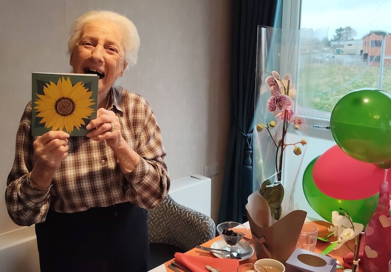 elderly lady sat at the table smiling holding up a photo of a painted sunflower